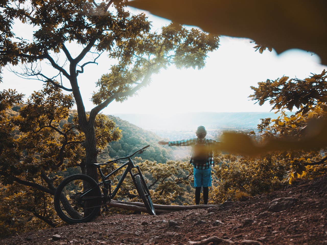 who-we-are A person stands with arms outstretched beside a mountain bike, enjoying the view in an autumn forest.